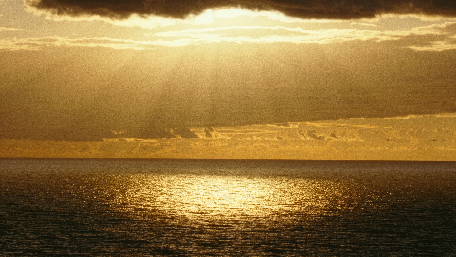 Golden light breaking through heavy clouds over a calm sea, rays fanning down onto the water