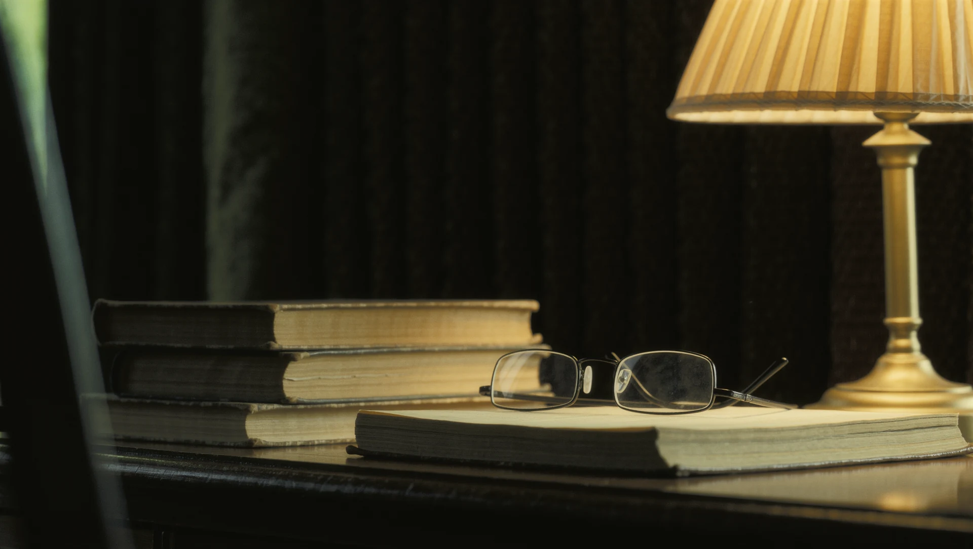 A stack of well-worn books on a desk beside reading glasses and a warm lamp, dark room, light pooling on the pages