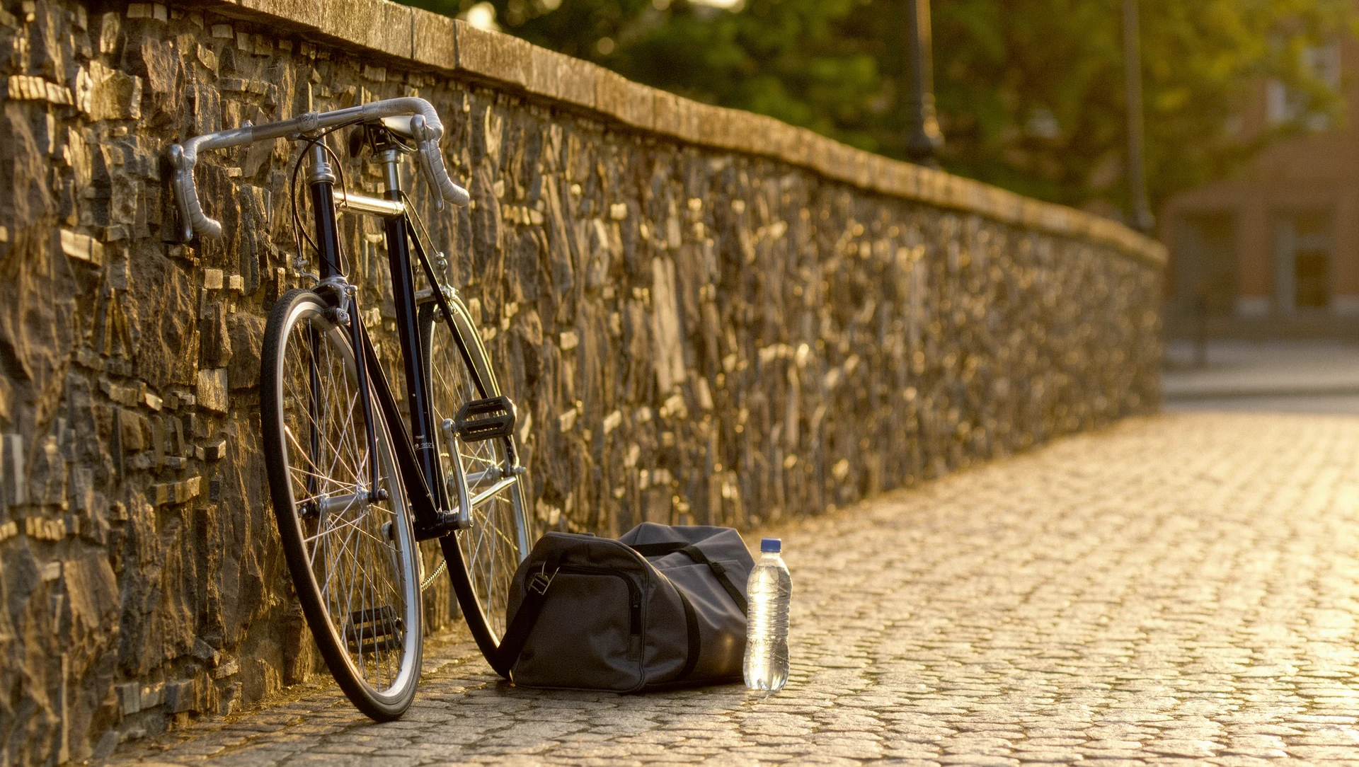 A bicycle leaning against a stone wall beside a packed gym bag and a water bottle, warm late afternoon sun