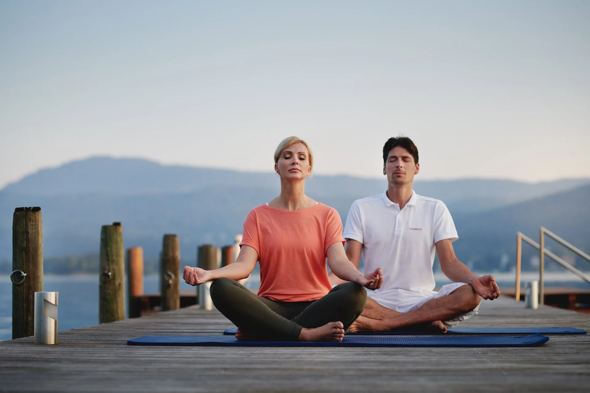 Two people meditating cross-legged on a VIVAMAYR wooden jetty by the lake