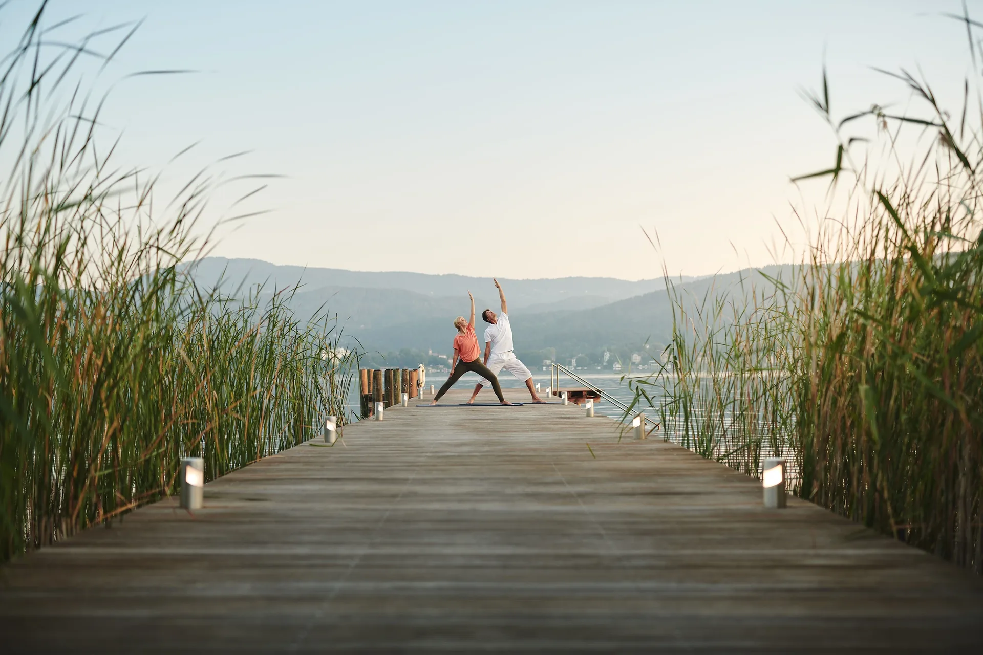 Two people doing yoga on a long wooden jetty between reeds, a VIVAMAYR Holistic Health Weeks scene