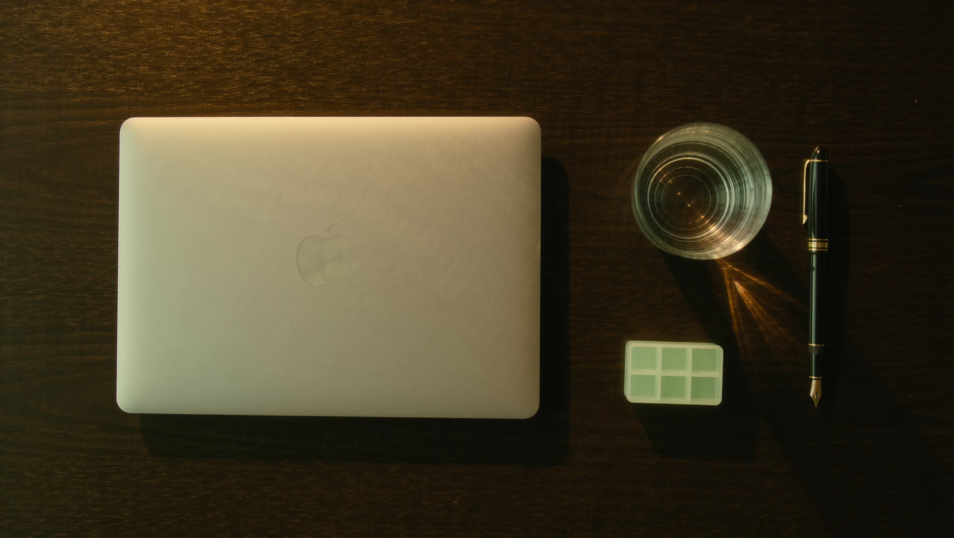 A minimalist workspace from above: a closed laptop, a glass of water, a small pill organiser and a fountain pen on dark wood