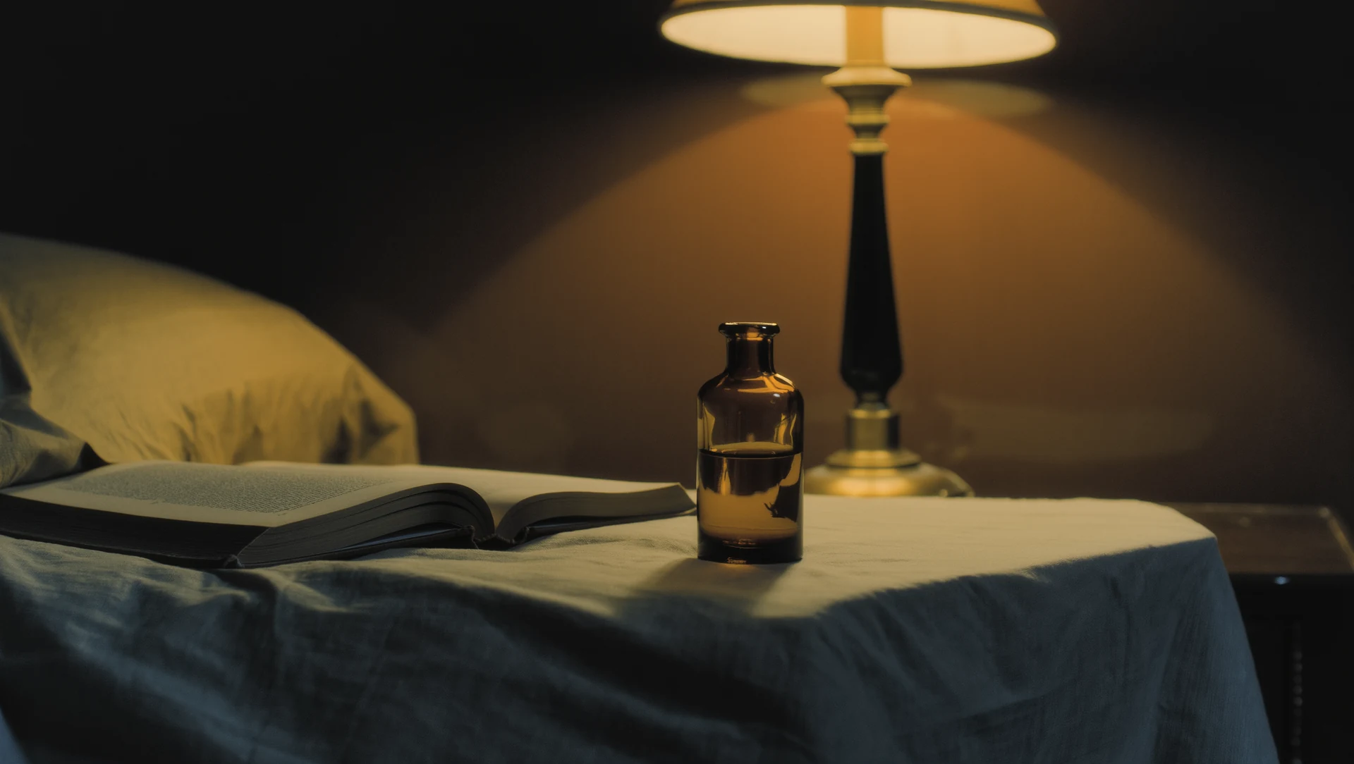 A bedside table at night: a small amber bottle, a book and a dimmed warm lamp, deep blue shadows across rumpled linen