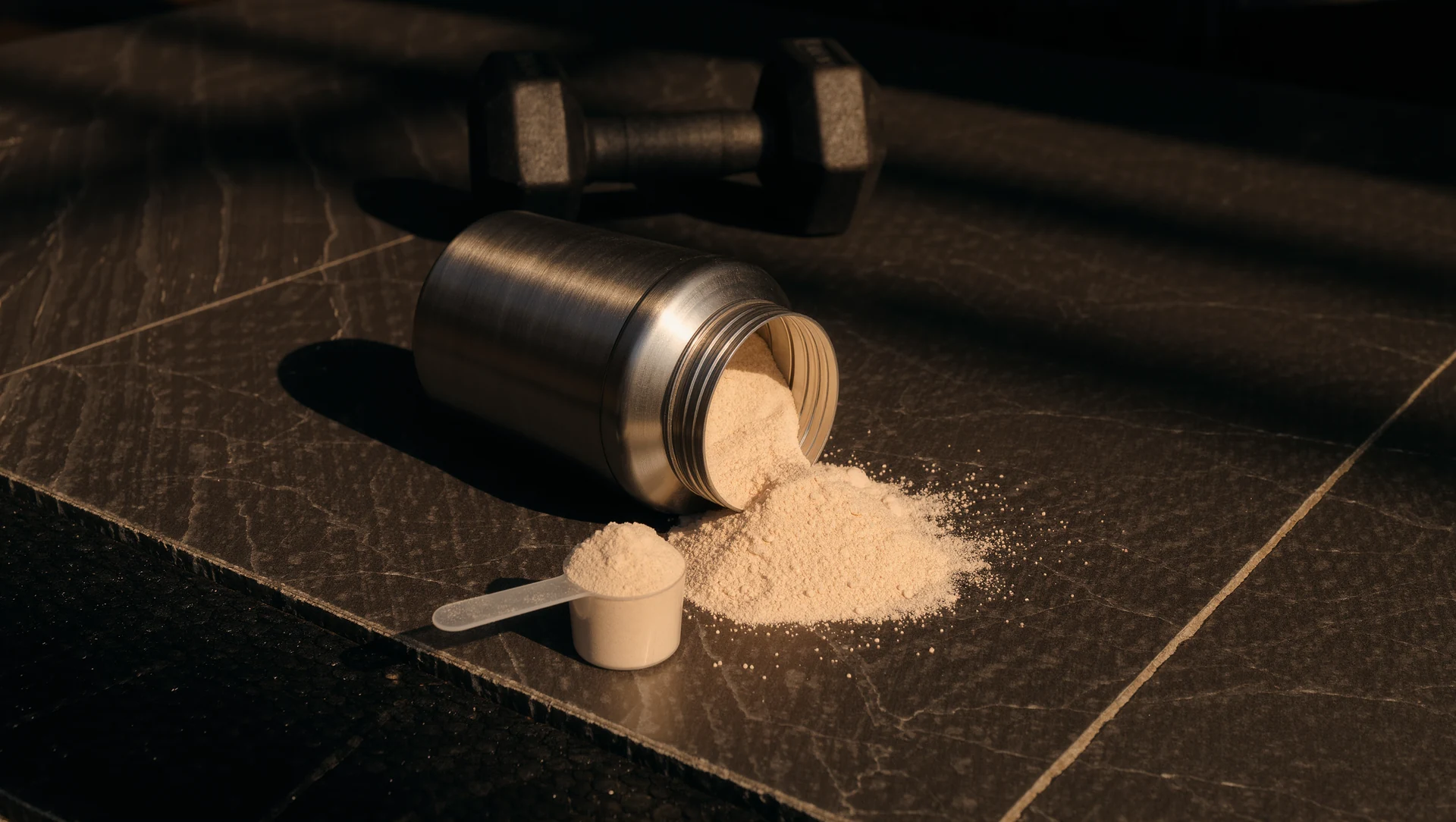 A steel shaker bottle, a scoop of protein powder spilling onto dark stone and a dumbbell just out of focus behind