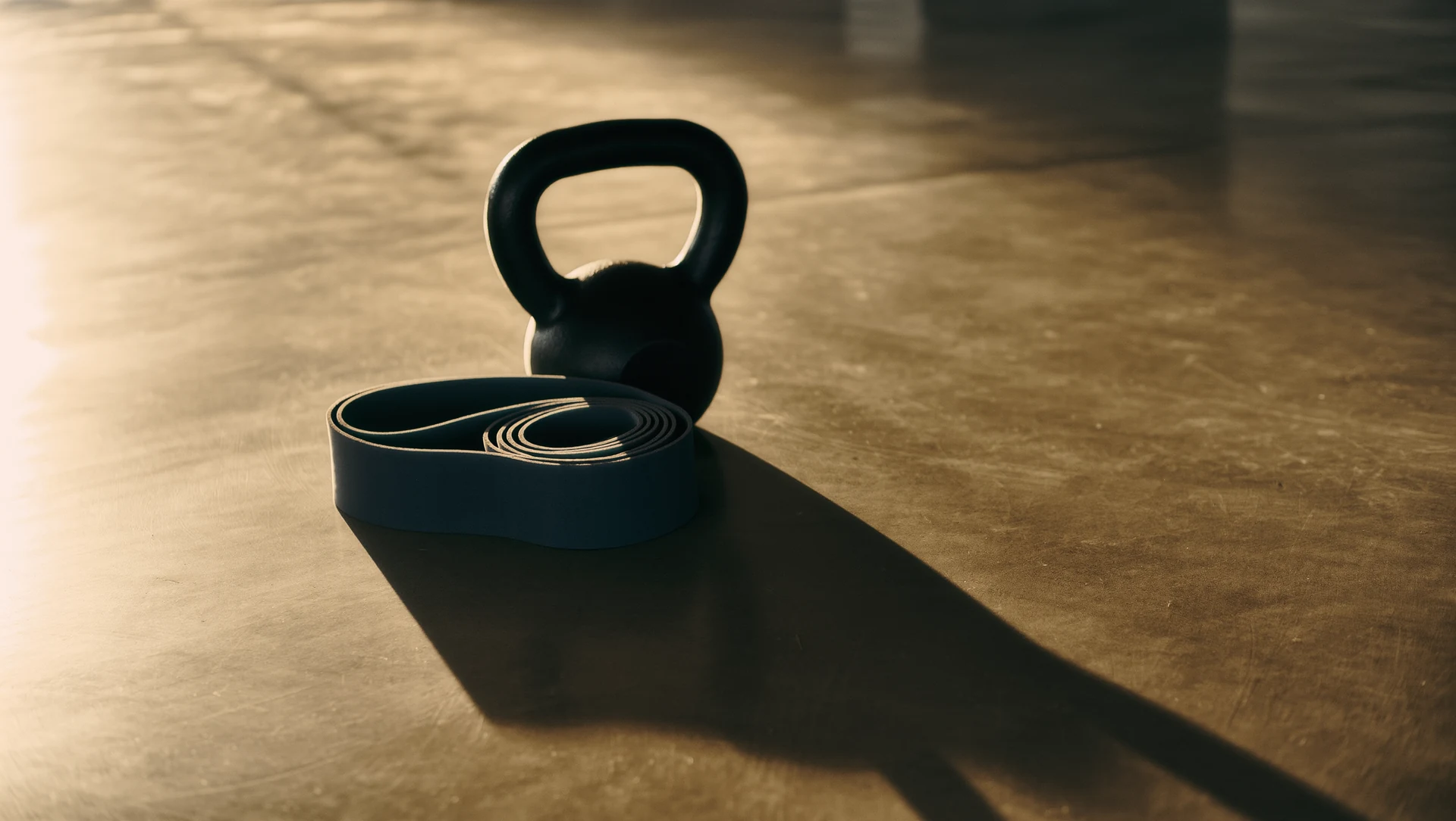 Resistance bands and a kettlebell arranged on polished concrete, long dramatic shadows from a single low light source