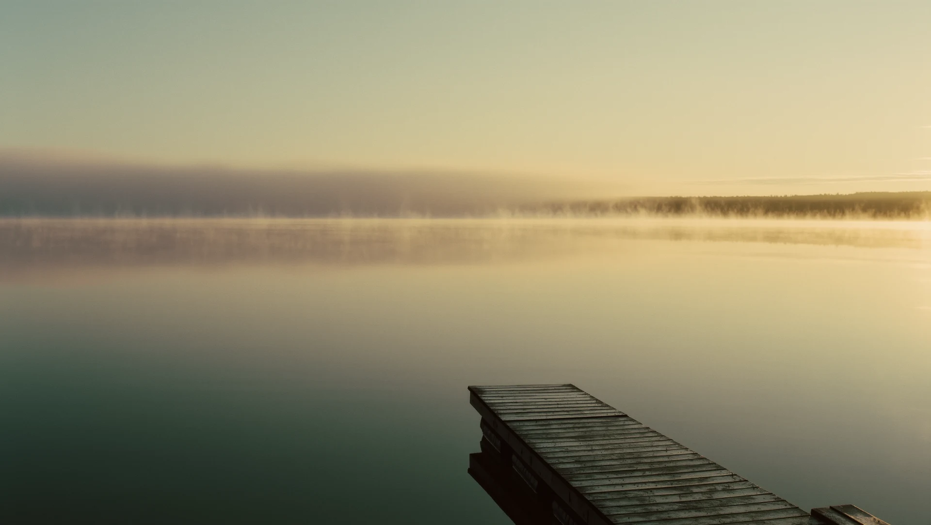 A still lake at dawn with mist on the surface and a single wooden jetty reaching into the water, warm light on the far shore