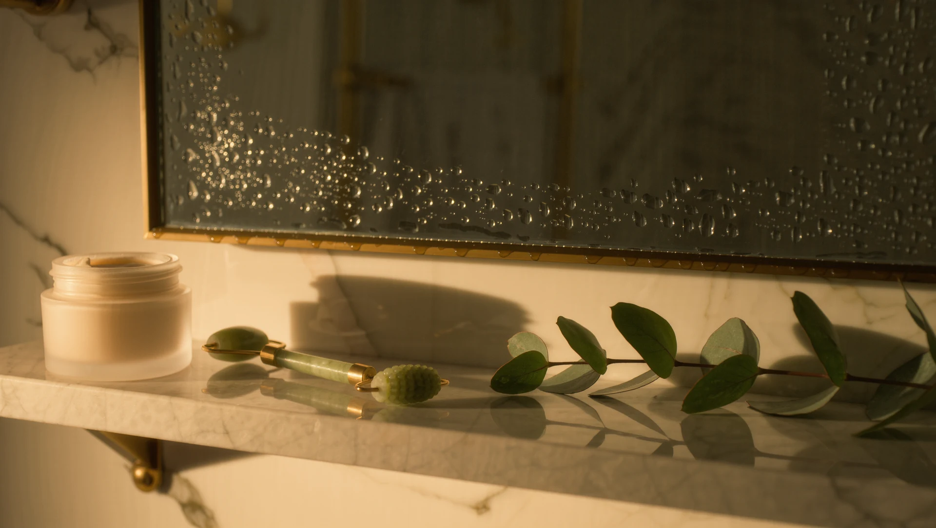 A marble bathroom shelf at dawn: a frosted glass jar of cream, a jade roller and a sprig of eucalyptus, condensation on a mirror behind