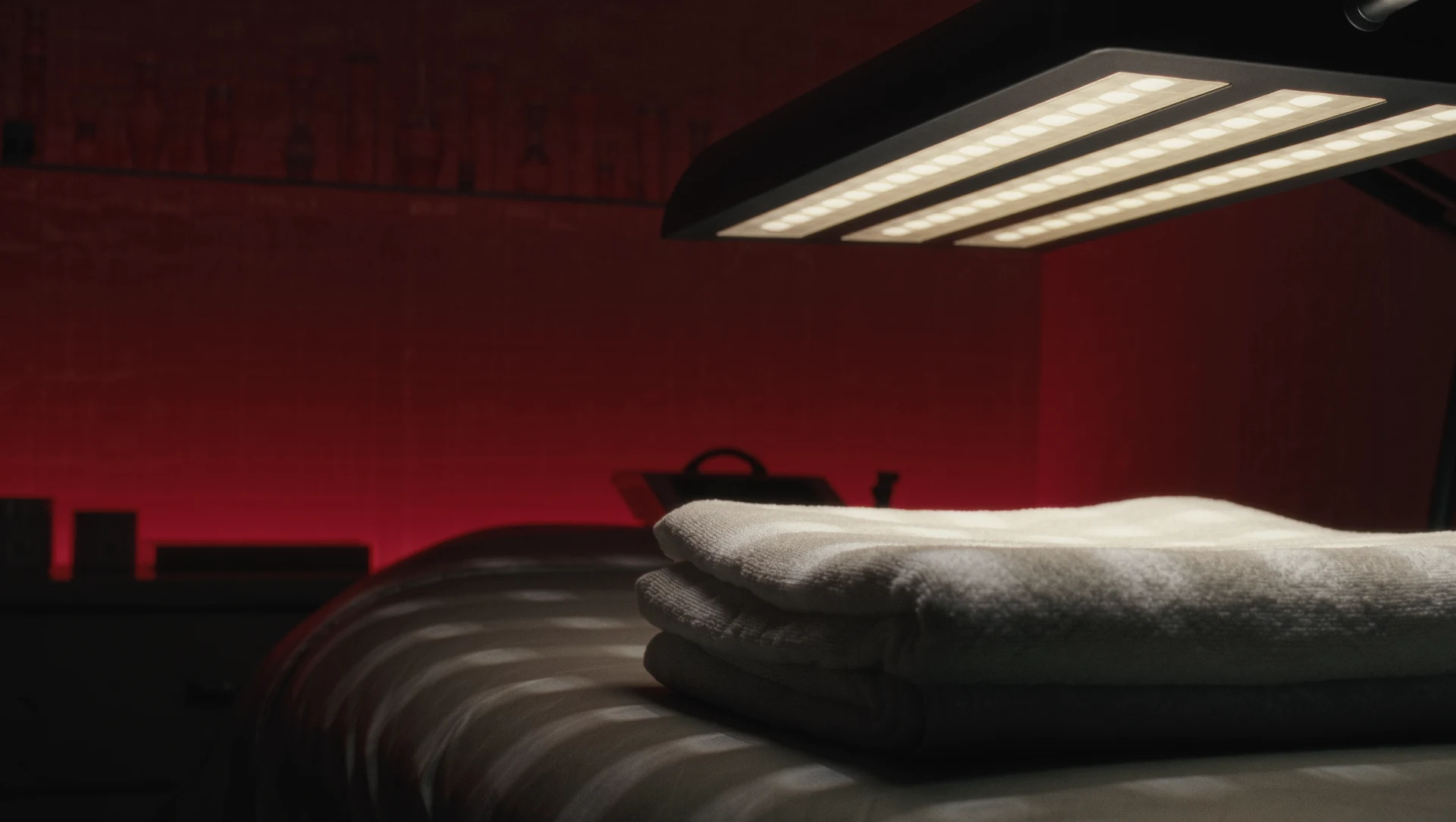 A darkened treatment room bathed in deep red light, a panel of soft glowing diodes casting warm patterns across a folded white towel