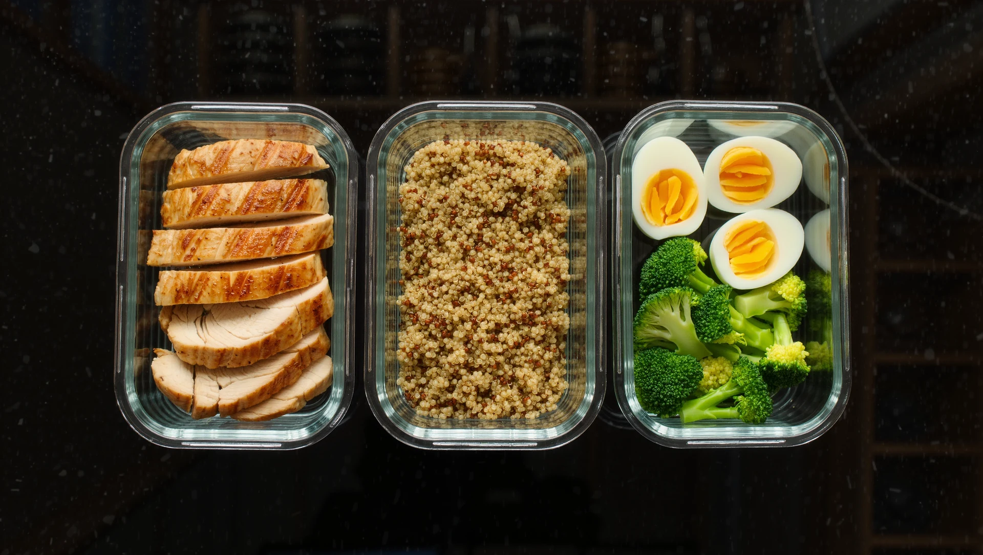 A meal-prep flatlay of glass containers with chicken, quinoa, eggs and steamed broccoli on a dark counter, warm overhead light