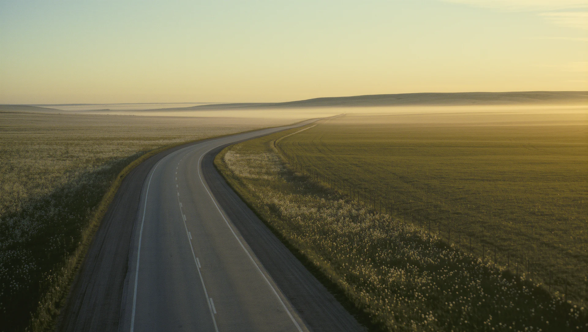 A long empty road curving through open countryside at golden hour, warm haze on the horizon